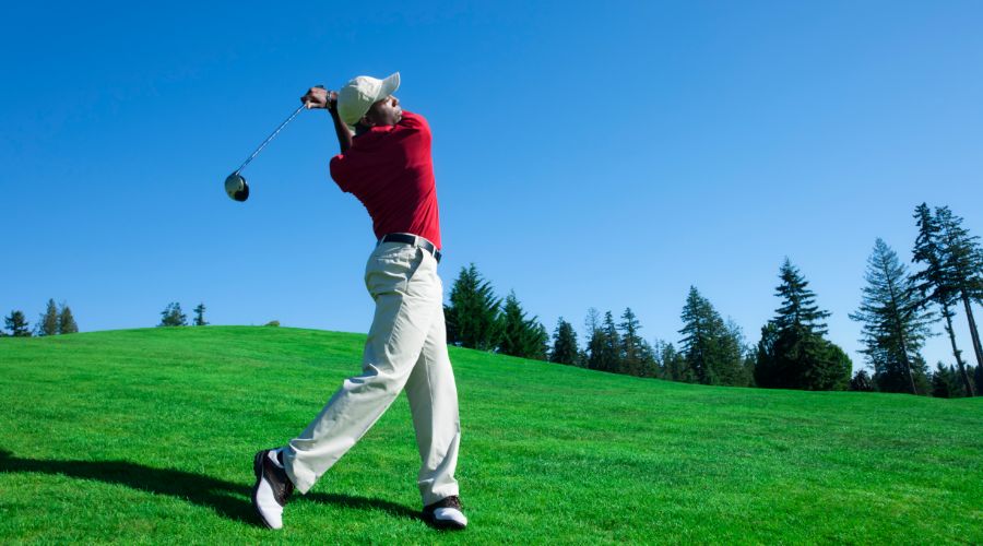 Golfer wearing a red shirt, beige pants, and a white cap in mid-swing on a lush green golf course surrounded by tall trees under a clear blue sky.